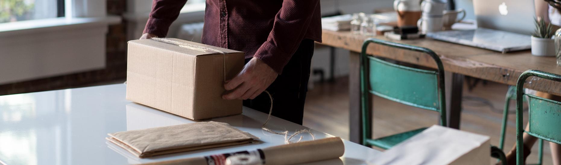 An Individual Wrapping Up Goods From Their Home Online Business, Using A Signet Cardboard Packaging Box