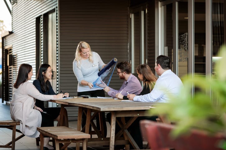 A group of Signet graduates seated in a covered area, observing a demonstration of pallet wrapping techniques by a blonde woman.