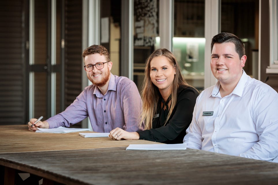 Three Signet graduates wearing business attire, sitting at a wooden table smiling at the camera